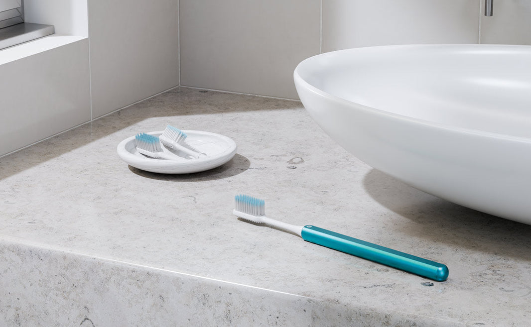 Arctic Blue Nada Toothbrush sitting on a bathroom counter with two brush heads in a dish 
