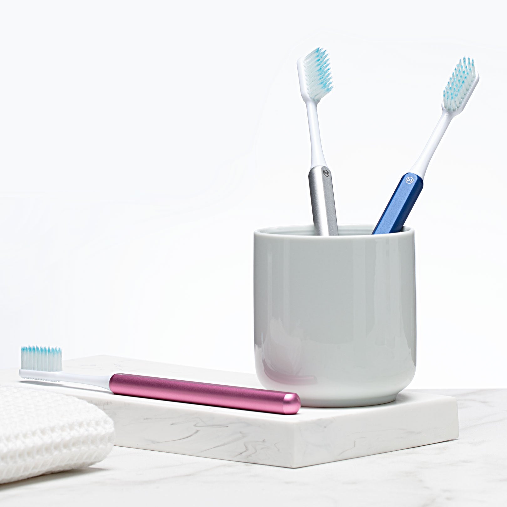Pink, Silver and Ocean blue Nada toothbrushes on a counter with a cup and a towel